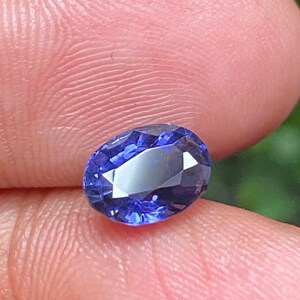 May include: Oval-shaped, deep blue gemstone with a faceted cut. The gemstone is set against a light pink background.