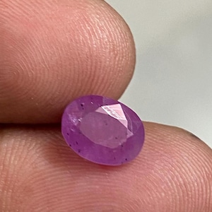 May include: A faceted, oval-shaped, purplish-pink gemstone. The gem is held between two fingers, showcasing its polished surfaces and internal clarity. The stone's color is a vibrant, saturated hue, reflecting light across its many facets.