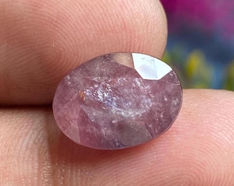 May include: A faceted, oval-shaped gemstone with a soft, purplish-pink hue. The gem has internal inclusions and a polished surface, reflecting light. The stone is held between fingers, showcasing its size and color.