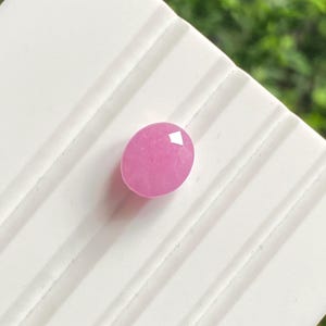 Pode incluir: Uma pedra preciosa rosa facetada em forma oval. A gema está colocada sobre uma superfície branca e canelada, com um fundo verde desfocado. As facetas da gema capturam a luz, criando um efeito brilhante. A gema é de um tom rosa vibrante.