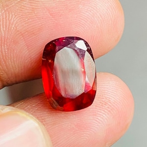 May include: A red gemstone with a faceted cut. The gemstone is held in a hand.