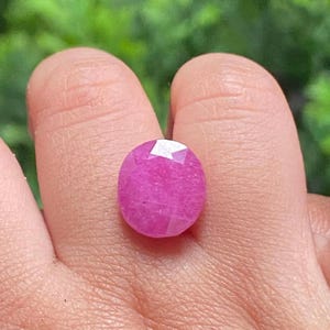 Pode incluir: Uma pedra preciosa rosa facetada, em forma oval. A gema é segurada em uma mão, com um fundo verde desfocado. A pedra preciosa tem uma superfície polida com múltiplas facetas que refletem a luz. A cor é um rosa vibrante e saturado.