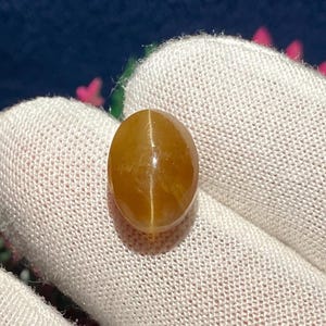 May include: A golden-brown, oval-shaped gemstone with a cat's eye effect, resting on a white fabric. The gemstone has a smooth, polished surface and a subtle, internal line of light. The background is a blurred mix of blue and pink.