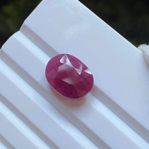 Puede incluir: Una gema de rubí facetada, de forma ovalada. El rubí es de un color rojo intenso y se encuentra sobre un fondo blanco. La gema tiene una superficie pulida con facetas visibles.