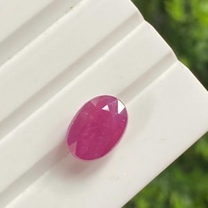 May include: A faceted, oval ruby gemstone. The gem is a deep, saturated pinkish-red colour, with visible facets that catch the light. The ruby is set against a white, ridged surface, with a blurred green background.
