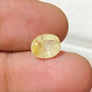 Puede incluir: Una piedra preciosa ovalada de color amarillo con un corte facetado. La piedra preciosa tiene una apariencia translúcida con un ligero brillo.