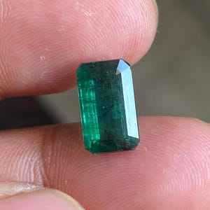 May include: A deep green emerald gemstone cut into a rectangular shape. The emerald is faceted and has a polished finish.
