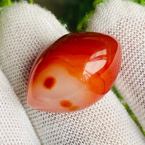 May include: A polished, teardrop-shaped carnelian gemstone featuring a smooth, glossy surface with vibrant orange and deep red hues. The stone displays translucent layers and two distinct, darker circular inclusions within its translucent body.