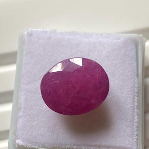 May include: A faceted, oval-shaped ruby gemstone, displayed in a white plastic case. The ruby has a deep pinkish-red colour and a polished surface. The case is set against a light background.
