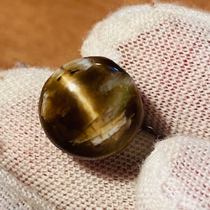 May include: A round, polished gemstone featuring a chatoyant cat's eye effect with a prominent vertical band of light. The stone displays a deep brown base color with translucent, cloudy white and cream-colored inclusions throughout its surface.