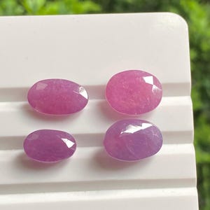May include: Four oval-shaped, faceted gemstones in varying shades of pink and purple. The gems are displayed on a white, ridged surface, with a blurred green background.