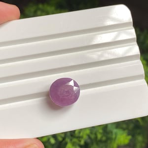 May include: A round, faceted, purplish-pink gemstone sits on a white, grooved surface. The gem's cut displays a star-like pattern. The background is blurred green foliage, contrasting with the gem's colour and the white surface.