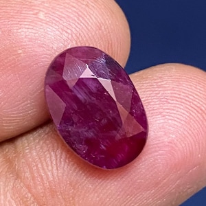 Puede incluir: Una piedra preciosa ovalada de color rojo intenso con un corte facetado. La piedra preciosa tiene una apariencia ligeramente translúcida y muestra algunas inclusiones internas.