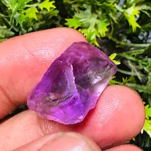 Pode incluir: Um cristal de ametista roxo com uma textura áspera e natural. O cristal é translúcido e tem um tom roxo claro.