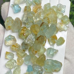 May include: A collection of raw aquamarine gemstones in various shapes and sizes, with colors ranging from pale blue to yellow-green. The gemstones are displayed on a white tray, showcasing their natural, unpolished state.