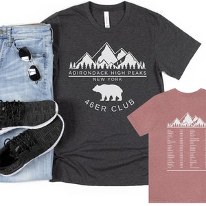 May include: Two t-shirts with a white graphic design of mountains, trees, and a bear. The text on the shirts reads "Adirondack High Peaks New York 46er Club". The shirt on the left is dark heather grey and the shirt on the right is a light pink.