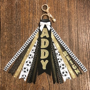 May include: Black and gold soccer-themed keychain with the name "ADDY" in gold glitter letters. The keychain has a white bow and multiple ribbons with black and white soccer balls, black and white chevron, and gold glitter.