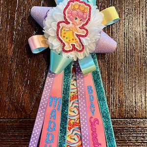 May include: A decorative birthday badge featuring a central white ruffled rosette with a cartoon character wearing a yellow dress and pink hair. The badge has layered ribbons in light blue, lavender, and yellow, with hanging streamers reading MADDY and BDAY Sweetie.
