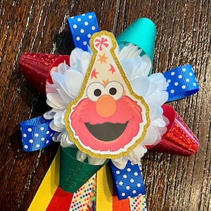 May include: A decorative rosette featuring a central cutout of a red Elmo character wearing a party hat with gold glitter trim. The center is surrounded by white fabric petals and layered with multi-colored ribbons in blue, red, green, and yellow.