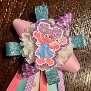 May include: A decorative hair bow featuring a central cutout of Abby Cadabby with pink skin, blue dress, and wings. The bow is constructed from layers of light blue glitter ribbon, pink ribbon, purple patterned ribbon, and white ruffled fabric.