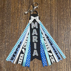 May include: A black and silver cheer keychain with the name "MARIA" on it. The keychain has blue, white, and silver ribbons with the words "cheer" and "cheerleader" on them.