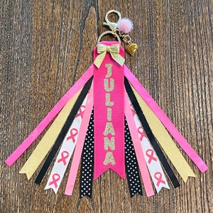 May include: Pink and gold ribbon keychain with the name "JULIANA" on it. The keychain has a pink bow and a small gold heart charm.