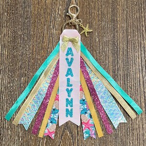 May include: A pink ribbon keychain with the name "AVALynn" in turquoise glitter letters. The keychain has a gold star charm and multiple colorful ribbons, including turquoise, gold, iridescent, and pink.