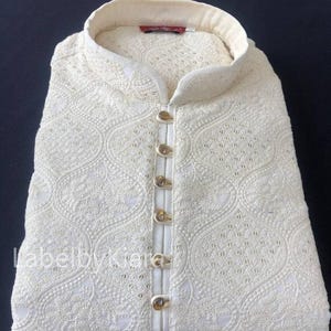May include: A white cotton kurta with intricate embroidery and gold button closures. The kurta has a mandarin collar and a long, flowing silhouette.