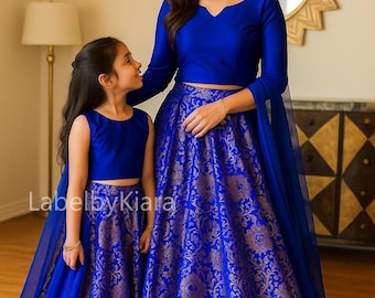 May include: Matching blue outfits. A woman and a child are wearing coordinating blue crop tops and long skirts with gold floral patterns. The woman's outfit has long sleeves, while the child's is sleeveless. The outfits are from LabelbyKiara.
