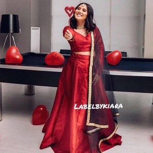 May include: A woman wearing a red crop top and a long red skirt with a black sheer dupatta with gold trim. She is holding a red heart-shaped wand and smiling. The text "LABELBYKIARA" is visible on the skirt.