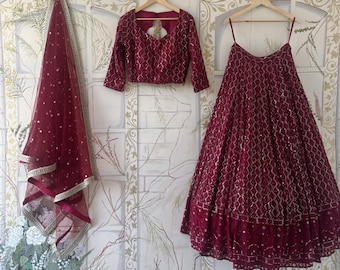 May include: A maroon bridal lehenga set with a matching embroidered blouse and a sheer dupatta. The skirt is embellished with silver sequins and the blouse has a deep V-neckline.