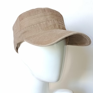 Peut inclure: Casquette de style militaire de couleur beige avec une visière plate et une couronne structurée. La casquette présente des coutures horizontales sur le dessus et un aspect légèrement vieilli. Le chapeau est présenté sur une tête de mannequin blanche.