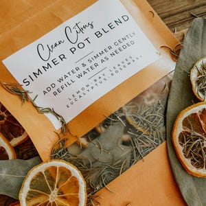 May include: An orange paper packet labeled "Clean Citrus SIMMER POT BLEND" with a clear window showing dried herbs. The packet is surrounded by dried lemon slices, rosemary sprigs, and eucalyptus leaves. The text on the label reads "Add water & simmer gently, refill water as needed."