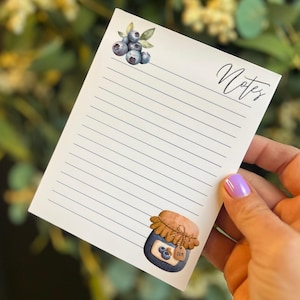 May include: A white notepad with lined pages, featuring watercolor illustrations of blueberries and a jam jar. The word "Notes" is written in cursive. The notepad is held by a person with a purple nail polish.