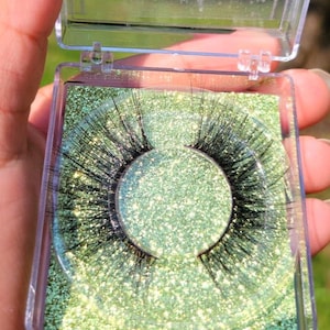 May include: A pair of false eyelashes in a clear plastic case with a green glitter background.