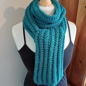 May include: A teal crochet scarf featuring a thick, ribbed texture. The scarf is wrapped around the neck of a mannequin, displaying a long, rectangular shape with vertical stitch patterns that create a consistent, raised appearance throughout the piece.