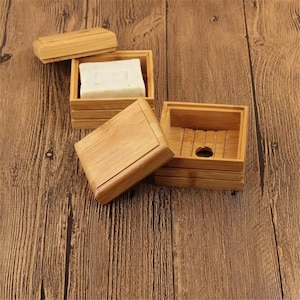 May include: A set of three square bamboo soap dishes. The top dish has a lid and the bottom dish has a drainage hole. The middle dish is empty.