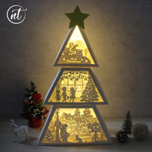 Peut inclure: Une lumière en forme de sapin de Noël blanche et superposée, avec une étoile verte au sommet. Chaque couche présente des scènes découpées complexes du Père Noël, d'enfants et d'un village, éclairées de l'intérieur. Le texte "Merry Christmas" est visible.