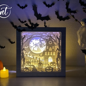 Puede incluir: Una caja blanca iluminada con una escena de Halloween. La escena incluye una casa encantada, una luna llena, murciélagos y un cementerio. La caja está iluminada desde el interior, creando un brillo cálido.