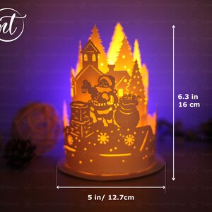 K&ouml;nnte beinhalten: Eine wei&szlig;e Weihnachts-Papierkunst-Szene mit einer Weihnachtsmann-Figur, die einen Sack mit Geschenken h&auml;lt. Die Szene wird von innen beleuchtet und erzeugt ein warmes Leuchten. Die Szene ist 16 cm hoch und 12,7 cm breit.