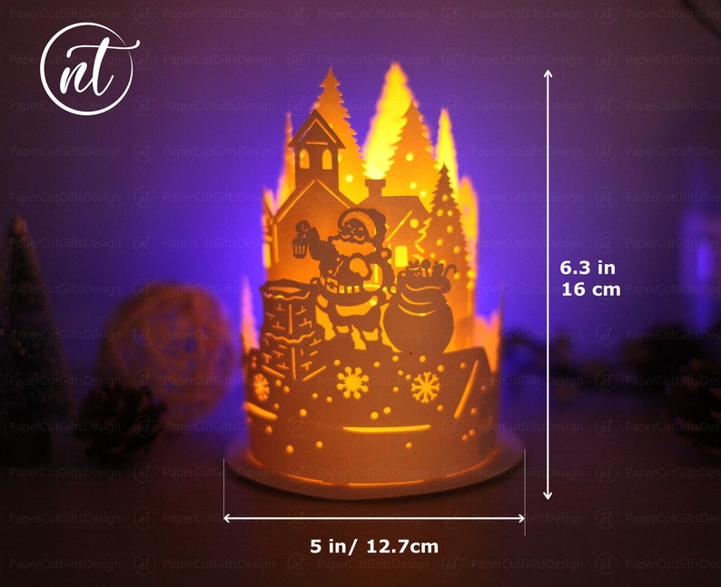 K&ouml;nnte beinhalten: Eine wei&szlig;e Weihnachts-Papierkunst-Szene mit einer Weihnachtsmann-Figur, die einen Sack mit Geschenken h&auml;lt. Die Szene wird von innen beleuchtet und erzeugt ein warmes Leuchten. Die Szene ist 16 cm hoch und 12,7 cm breit.