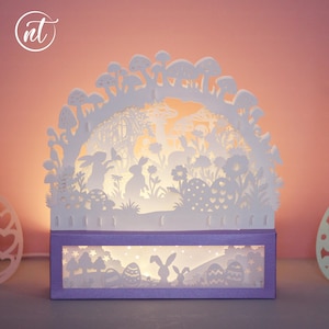 Puede incluir: Decoración luminosa de Pascua de papel blanco. El diseño presenta siluetas de conejos, huevos, flores y setas. La base es una caja rectangular con un borde morado. La luz brilla a través de los detalles recortados, creando un brillo cálido.