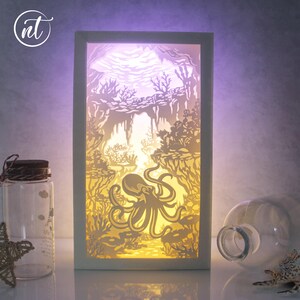 May include: A white wooden lamp with a cutout design of an octopus in a coral reef. The lamp is lit from behind, creating a warm glow.