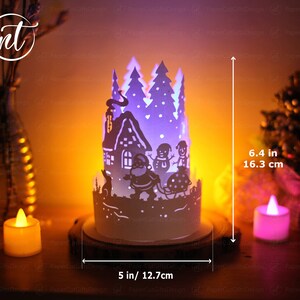 K&ouml;nnte beinhalten: Eine wei&szlig;e Weihnachts-Papierkunst mit einem Haus, B&auml;umen und Schneem&auml;nnern. Die Szene wird von innen beleuchtet und erzeugt ein warmes Leuchten. Die Szene ist 16,3 cm hoch und 12,7 cm breit.