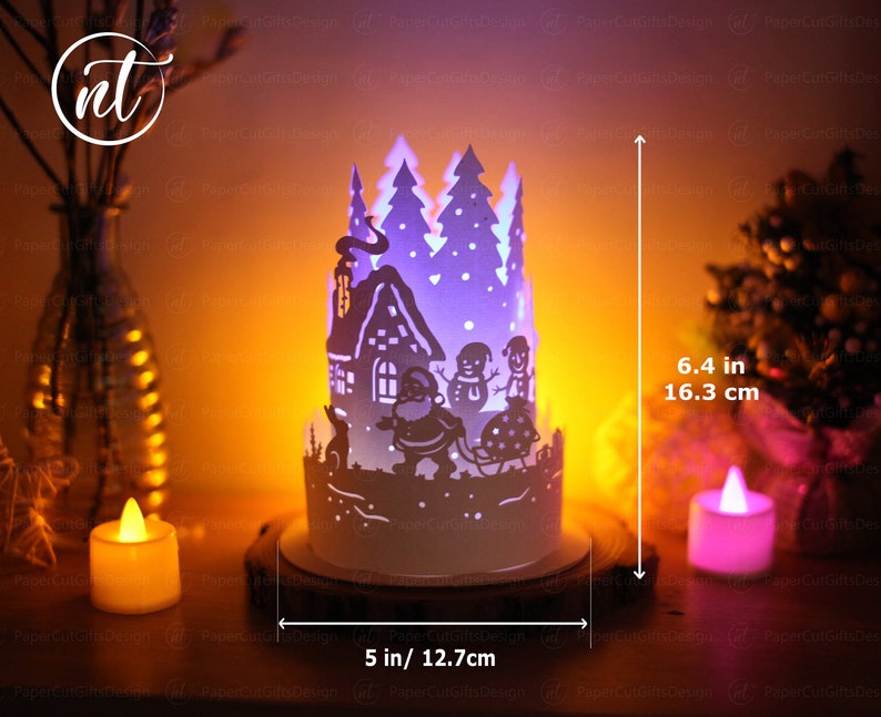 K&ouml;nnte beinhalten: Eine wei&szlig;e Weihnachts-Papierkunst mit einem Haus, B&auml;umen und Schneem&auml;nnern. Die Szene wird von innen beleuchtet und erzeugt ein warmes Leuchten. Die Szene ist 16,3 cm hoch und 12,7 cm breit.