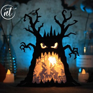 Op de afbeelding: Een zwarte papieren Halloween-lantaarn in de vorm van een boom met een griezelig gezicht. De lantaarn heeft een lichtje erin, dat een sc&egrave;ne met skeletten en heksen verlicht.