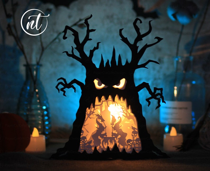 Op de afbeelding: Een zwarte papieren Halloween-lantaarn in de vorm van een boom met een griezelig gezicht. De lantaarn heeft een lichtje erin, dat een sc&egrave;ne met skeletten en heksen verlicht.