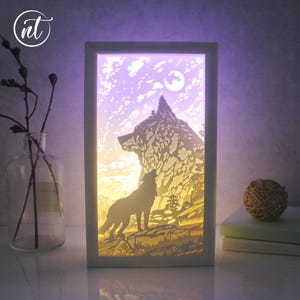 May include: A framed, layered paper art piece depicting a wolf howling at the moon. The artwork features a mountain silhouette, trees, and a starry sky, illuminated with a soft, warm glow. The frame is white.