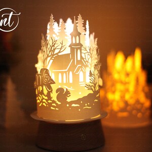 K&ouml;nnte beinhalten: Eine wei&szlig;e Papierlampe mit einem ausgeschnittenen Design einer Kirche in einem verschneiten Wald. Die Lampe ist beleuchtet und strahlt ein warmes Licht aus. Die Lampe steht auf einem Holzsockel.