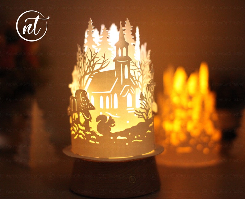 K&ouml;nnte beinhalten: Eine wei&szlig;e Papierlampe mit einem ausgeschnittenen Design einer Kirche in einem verschneiten Wald. Die Lampe ist beleuchtet und strahlt ein warmes Licht aus. Die Lampe steht auf einem Holzsockel.